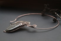 An elegant necklace with a unique design featuring a large, polished stone pendant. The stone is encased in intricate wirework that forms swirls and loops, giving it a sophisticated and artistic appearance. The necklace is displayed on a dark surface, and there are blurred floral elements in the background.