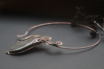 An elegant necklace with a unique design featuring a large, polished stone pendant. The stone is encased in intricate wirework that forms swirls and loops, giving it a sophisticated and artistic appearance. The necklace is displayed on a dark surface, and there are blurred floral elements in the background.