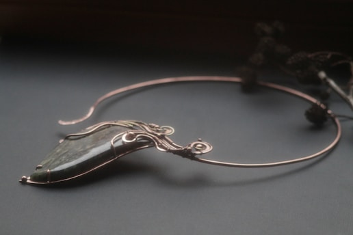 An elegant necklace with a unique design featuring a large, polished stone pendant. The stone is encased in intricate wirework that forms swirls and loops, giving it a sophisticated and artistic appearance. The necklace is displayed on a dark surface, and there are blurred floral elements in the background.
