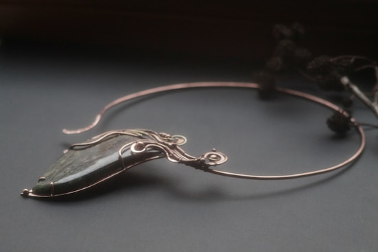 An elegant necklace with a unique design featuring a large, polished stone pendant. The stone is encased in intricate wirework that forms swirls and loops, giving it a sophisticated and artistic appearance. The necklace is displayed on a dark surface, and there are blurred floral elements in the background.
