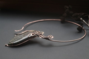 An elegant necklace with a unique design featuring a large, polished stone pendant. The stone is encased in intricate wirework that forms swirls and loops, giving it a sophisticated and artistic appearance. The necklace is displayed on a dark surface, and there are blurred floral elements in the background.