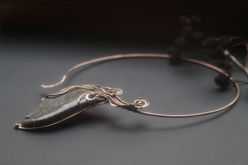 An elegant necklace with a unique design featuring a large, polished stone pendant. The stone is encased in intricate wirework that forms swirls and loops, giving it a sophisticated and artistic appearance. The necklace is displayed on a dark surface, and there are blurred floral elements in the background.