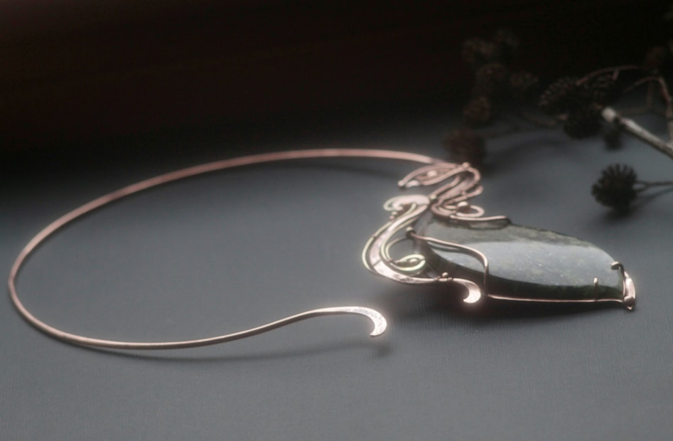 An elegant necklace with an intricately designed metal setting holding a polished, dark green stone. The setting features delicate, flowing curves, and the background shows some blurred out foliage.