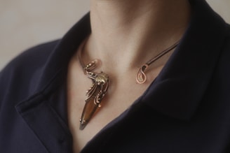 A close-up of a person's neck wearing an artistic necklace with a brown gem and intricate metallic design, set against a dark fabric background.