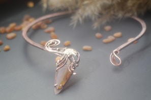 A delicate wire-wrapped necklace featuring an elongated gemstone in the center with artful metalwork curving around it. The background includes several scattered small stones and a soft, blurred texture adding to the aesthetic charm.