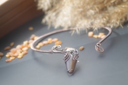 A delicate, intricately designed necklace with a teardrop-shaped gemstone pendant lies on a surface. The pendant is encased in a decorative metal setting, featuring ornate swirls. Surrounding the necklace are small amber-colored stones scattered across the surface and a textured golden material in the background.