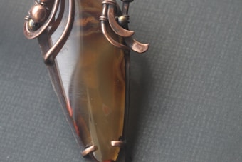 An intricately designed copper wire-wrapped pendant encasing a polished brown gemstone. The copper wires are artistically twisted around the top of the stone, adding an ornamental and handcrafted look.