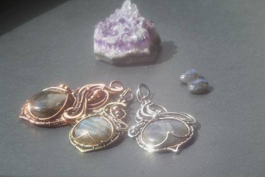 A trio of intricately crafted pendants featuring circular stones encased in metal wire settings, with varied metallic finishes of gold, copper, and silver. They are positioned on a dark surface alongside a small cluster of raw amethyst crystal and two small polished stones.