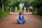 A person in a blue outfit sits cross-legged in a meditative pose on a dirt pathway lined with green foliage. The background features stone structures and trees, creating a serene, natural environment.