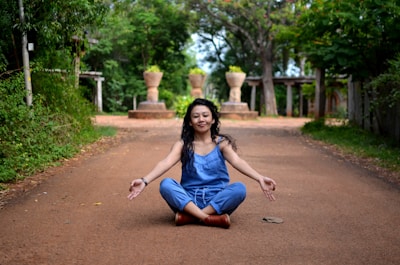 A person in a blue outfit sits cross-legged in a meditative pose on a dirt pathway lined with green foliage. The background features stone structures and trees, creating a serene, natural environment.