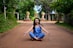A person in a blue outfit sits cross-legged in a meditative pose on a dirt pathway lined with green foliage. The background features stone structures and trees, creating a serene, natural environment.