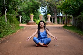 A person in a blue outfit sits cross-legged in a meditative pose on a dirt pathway lined with green foliage. The background features stone structures and trees, creating a serene, natural environment.