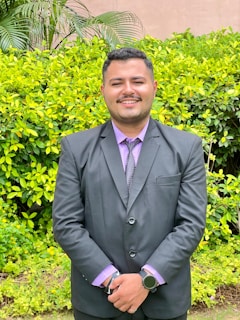 A person in a formal black suit with a purple shirt stands smiling in front of a lush green hedge. The setting appears to be outdoors with bright foliage and a few tall plants visible in the background.