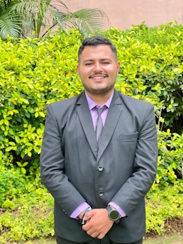 A person in a formal black suit with a purple shirt stands smiling in front of a lush green hedge. The setting appears to be outdoors with bright foliage and a few tall plants visible in the background.