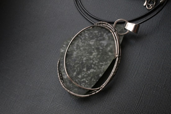 A delicate pendant featuring a polished, oval-shaped stone with a speckled pattern. The stone is secured in an intricately wrapped wire setting, attached to a black leather cord. The pendant's metallic elements have a silver finish, adding a touch of elegance.