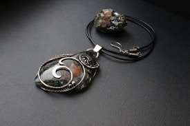 A pendant with intricate wire wrapping encases a large, dark stone. It is placed alongside a black cord necklace with a hook clasp. The stone displays a glossy surface with swirling wire patterns, and there is a rough, metallic rock in the background.