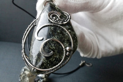 A hand wearing a white glove holds a pendant made of a dark, polished stone with intricate silver wire wrapping and decorative spirals. The background is a soft, blurred grey, and there is a small clip visible attached to the pendant.