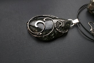 A teardrop-shaped pendant made from a dark green stone with intricate silver wire wrapping, placed on a black textured surface. The pendant is attached to a black cord, which is partially visible.