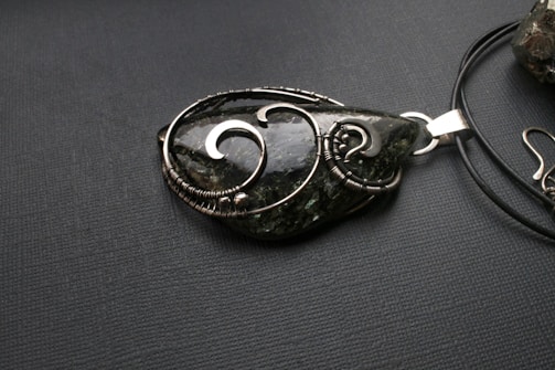 A teardrop-shaped pendant made from a dark green stone with intricate silver wire wrapping, placed on a black textured surface. The pendant is attached to a black cord, which is partially visible.