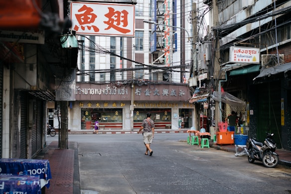 An urban street scene in an Asian city features a mix of older and newer architecture. Shop signs with Asian script hang above storefronts. A person walks down the relatively quiet street, lined with motorbikes, chairs, and signs of daily life. Overhead, numerous electrical cables stretch between buildings.