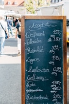 A blackboard menu with wooden frame displays a list of food items and their prices in German at an outdoor market. In the background, people are walking past various market stalls and food trucks under a sunny sky.