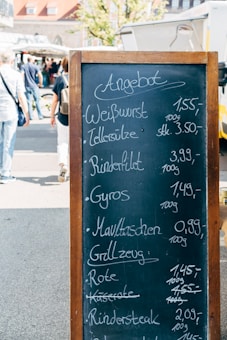 A blackboard menu with wooden frame displays a list of food items and their prices in German at an outdoor market. In the background, people are walking past various market stalls and food trucks under a sunny sky.