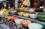 An assortment of colorful ingredients is displayed in glass bowls on a metal food stall counter. The bowls contain various items like chopped fruits, corn, beans, ice, jelly, and other dessert toppings. Packaged snacks are stacked in a basket nearby. A pricing label is visible, and a fan is attached to the stall. The setting appears to be a vibrant, bustling market.
