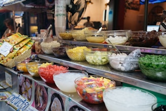 An assortment of colorful ingredients is displayed in glass bowls on a metal food stall counter. The bowls contain various items like chopped fruits, corn, beans, ice, jelly, and other dessert toppings. Packaged snacks are stacked in a basket nearby. A pricing label is visible, and a fan is attached to the stall. The setting appears to be a vibrant, bustling market.