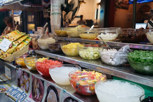 An assortment of colorful ingredients is displayed in glass bowls on a metal food stall counter. The bowls contain various items like chopped fruits, corn, beans, ice, jelly, and other dessert toppings. Packaged snacks are stacked in a basket nearby. A pricing label is visible, and a fan is attached to the stall. The setting appears to be a vibrant, bustling market.