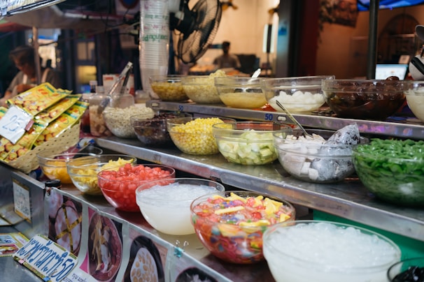 An assortment of colorful ingredients is displayed in glass bowls on a metal food stall counter. The bowls contain various items like chopped fruits, corn, beans, ice, jelly, and other dessert toppings. Packaged snacks are stacked in a basket nearby. A pricing label is visible, and a fan is attached to the stall. The setting appears to be a vibrant, bustling market.