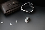 A black leather wallet with a bull logo embossed on it lies on a dark surface. Next to it, there is a silver and black wristwatch with a metal strap. A silver ring with a blue gemstone and other small oval gemstones are also arranged nearby.