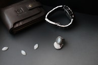 A black leather wallet with a bull logo embossed on it lies on a dark surface. Next to it, there is a silver and black wristwatch with a metal strap. A silver ring with a blue gemstone and other small oval gemstones are also arranged nearby.