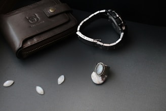 A black leather wallet with a bull logo embossed on it lies on a dark surface. Next to it, there is a silver and black wristwatch with a metal strap. A silver ring with a blue gemstone and other small oval gemstones are also arranged nearby.