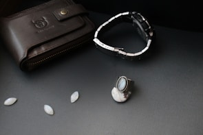 A black leather wallet with a bull logo embossed on it lies on a dark surface. Next to it, there is a silver and black wristwatch with a metal strap. A silver ring with a blue gemstone and other small oval gemstones are also arranged nearby.
