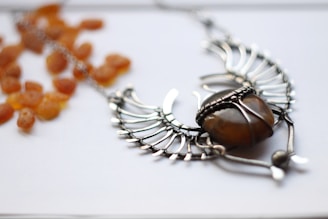 A silver, wire-wrapped necklace featuring an amber-colored stone, arranged in an artistic shape resembling wings or a bird. Lying beside the necklace are scattered dried apricot pieces, set against a white background.