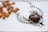 A silver, wire-wrapped necklace featuring an amber-colored stone, arranged in an artistic shape resembling wings or a bird. Lying beside the necklace are scattered dried apricot pieces, set against a white background.