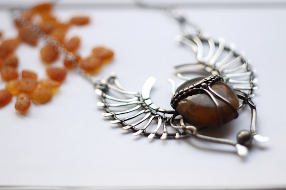 A silver, wire-wrapped necklace featuring an amber-colored stone, arranged in an artistic shape resembling wings or a bird. Lying beside the necklace are scattered dried apricot pieces, set against a white background.
