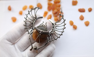 An intricately designed necklace pendant resembling a scarab beetle, featuring metallic wings and a polished amber stone at its center. The pendant is held by a white-gloved hand against a soft-focus background scattered with small amber stones.