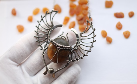 An intricately designed necklace pendant resembling a scarab beetle, featuring metallic wings and a polished amber stone at its center. The pendant is held by a white-gloved hand against a soft-focus background scattered with small amber stones.