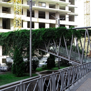 A metal pedestrian bridge covered with dense greenery is seen in front of a partially constructed building. The bridge, supported by steel beams, has a glass side and is surrounded by well-maintained bushes and small trees. In the background, several vehicles are parked near the construction site with visible scaffolding and unfinished concrete structures.