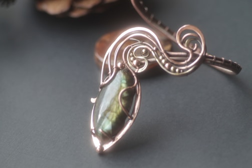 A beautifully detailed pendant featuring an intricately designed metal frame that loops and curves around an oval-shaped gemstone with a shiny, iridescent surface. The pendant rests on a flat, dark background, which highlights the craftsmanship and elegance of the piece.