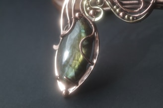 An intricately designed pendant featuring a polished, oval-shaped stone with an iridescent finish. The stone is encased in a decorative metal framework with ornate wire-wrapping and bead details.