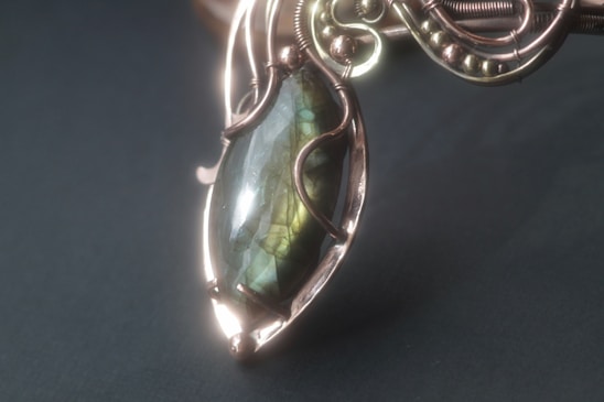 An intricately designed pendant featuring a polished, oval-shaped stone with an iridescent finish. The stone is encased in a decorative metal framework with ornate wire-wrapping and bead details.