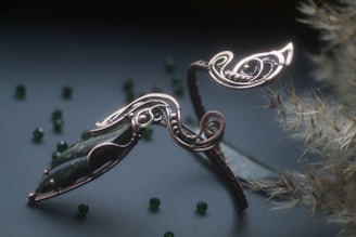 An intricately designed piece of metal jewelry, possibly a bracelet, featuring elegant curves and spirals. It has a dark stone centerpiece and is set against a textured background with scattered small green beads and pale dried plant elements.