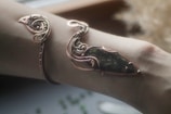 An intricately designed piece of jewelry wraps around an arm, featuring a mix of copper wire and a stone with dark green and black hues. The metalwork is elaborate, with swirling patterns and a glossy finish.
