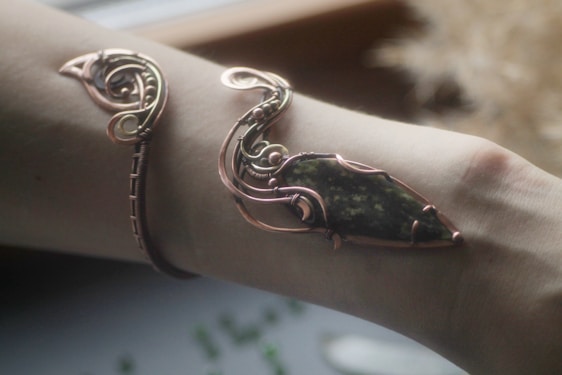An intricately designed piece of jewelry wraps around an arm, featuring a mix of copper wire and a stone with dark green and black hues. The metalwork is elaborate, with swirling patterns and a glossy finish.