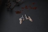 Two intricately designed pendants with tooth-like elements and gemstones are placed on a textured dark surface. The pendants are silver with one holding a blue gemstone and the other a black gemstone. Above them, there is a branch with pinecones and scattered reddish-brown powder, possibly cinnamon.