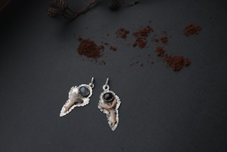 Two metallic earrings with intricate designs are placed on a dark textured surface. Each earring has a shiny, dark bead and what appears to be a small bone embellishment. Scattered around them is a sprinkling of brown powder. In the upper left corner, there is a dried branch with a few small cones attached.