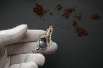 A white-gloved hand holds an ornate piece of jewelry with a blue stone and intricate metalwork. The background consists of a dark textured surface with scattered brown powder.