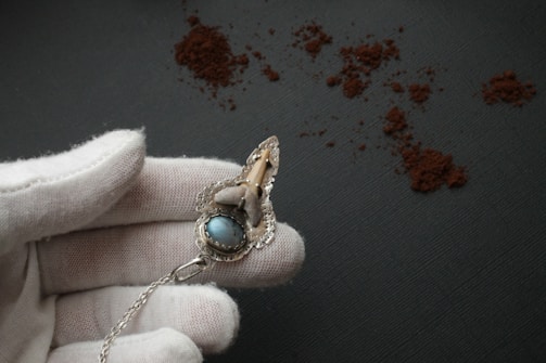 A white-gloved hand holds an ornate piece of jewelry with a blue stone and intricate metalwork. The background consists of a dark textured surface with scattered brown powder.